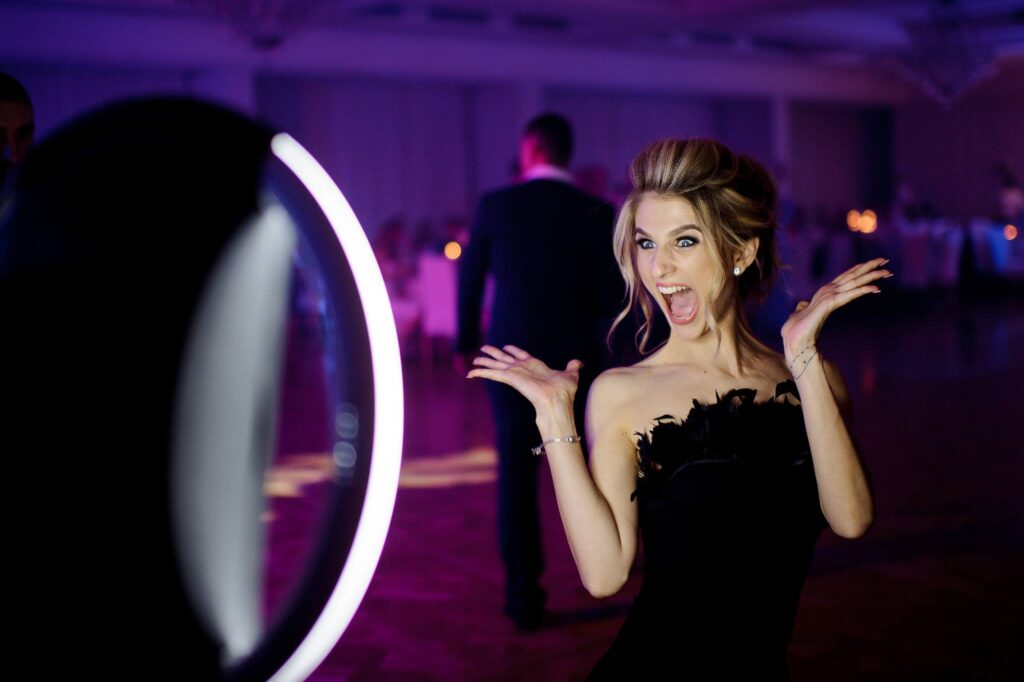 A woman in a black dress poses excitedly with her hands up in front of a ring light at an indoor event.