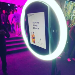 A photo booth with a ring light and a screen displaying "Tap to Start Booth" at an indoor event with purple lighting.