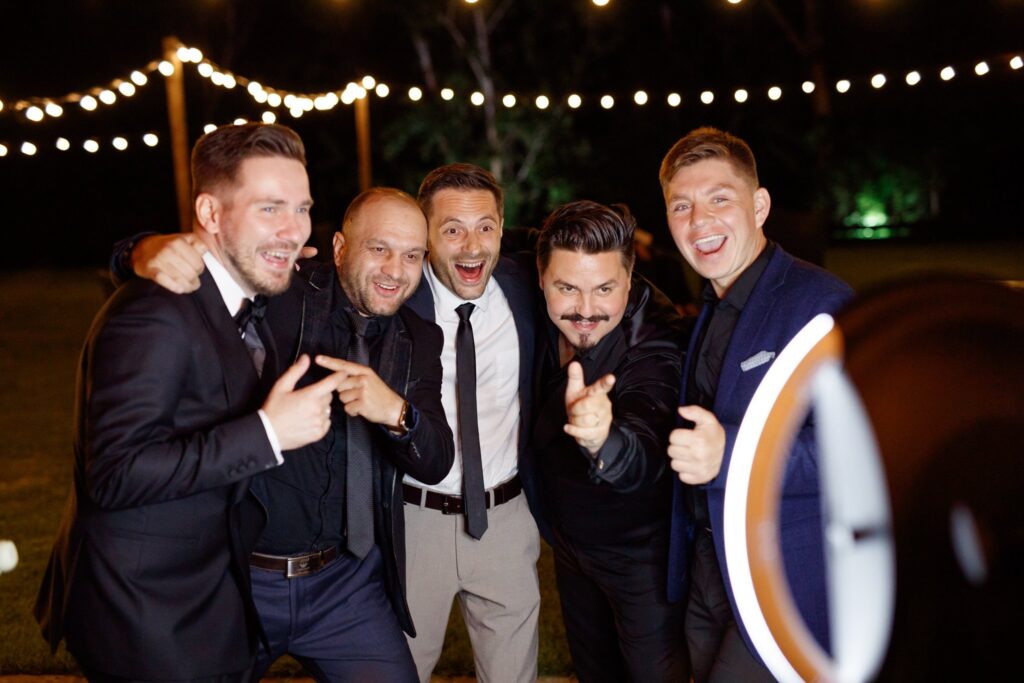 Five men in formal attire posing and laughing for a photo at an outdoor nighttime event with string lights in the background.