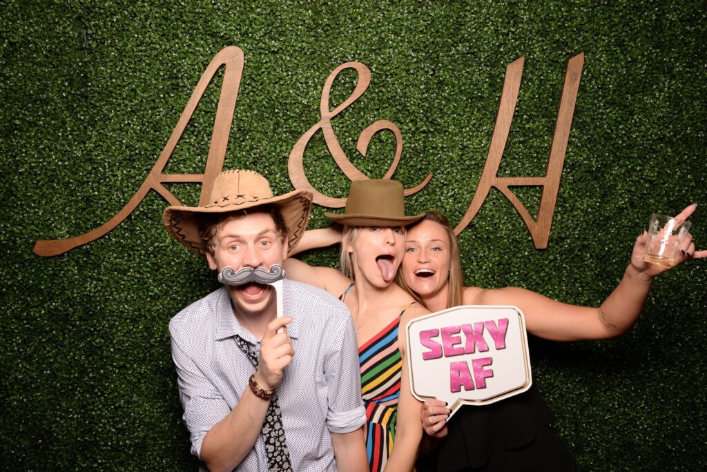 Three people pose in front of a leafy backdrop with the letters "A & H." They hold playful props and signs, with one person sticking out their tongue.