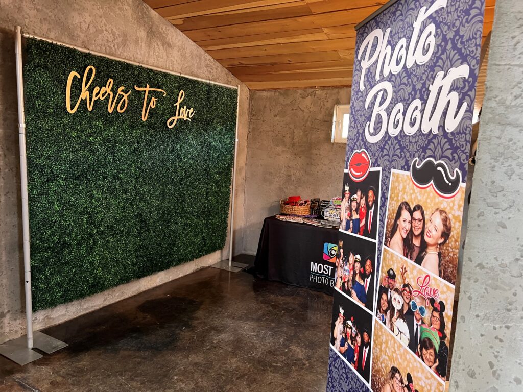 A photo booth setup with a green backdrop reading "Cheers to Love" and a collage of photos on the booth display.