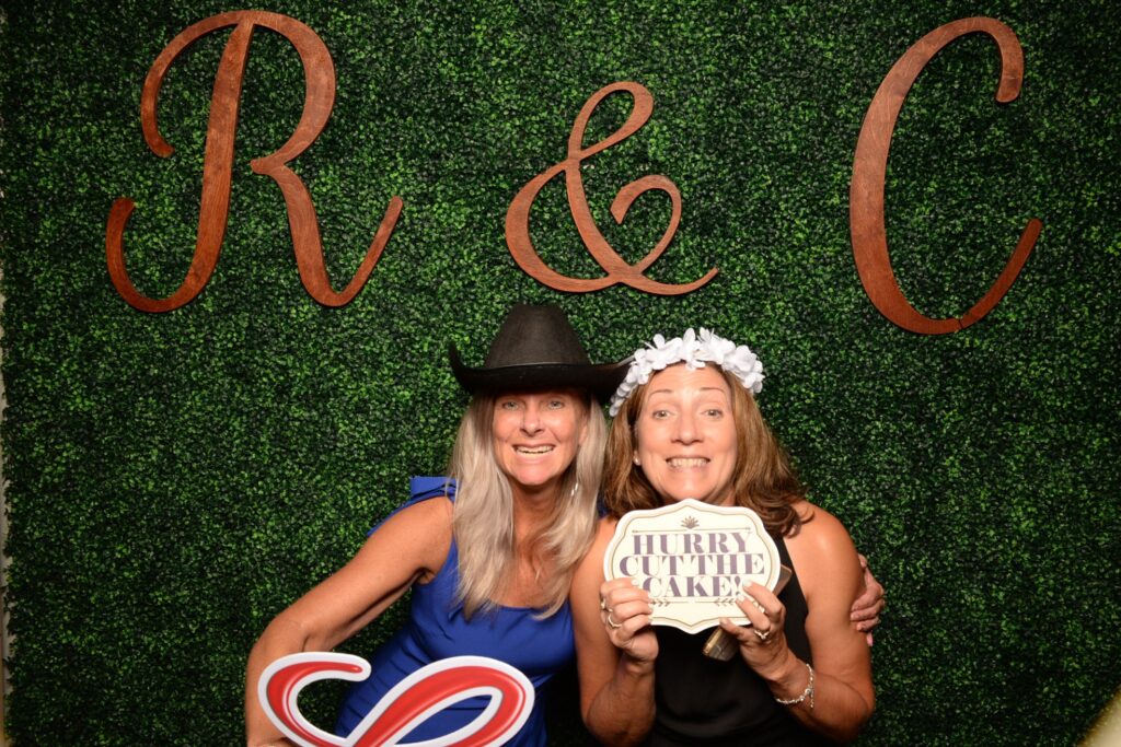 Two women pose in front of a greenery backdrop with large wooden letters "R & C"; one holds a heart sign, the other a "Hurry cut the cake" sign.