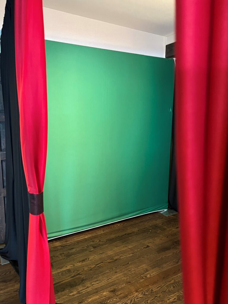 A green screen backdrop is set up in a room with hardwood flooring, flanked by red curtains on either side.