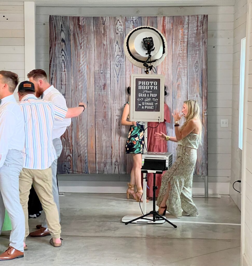 People using a photo booth with a wooden backdrop at an indoor event. A sign provides instructions to grab a prop, strike a pose.