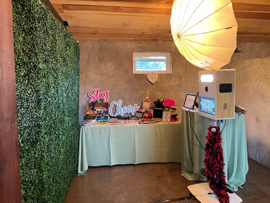 Photo booth setup with a green backdrop, props table, light umbrella, and camera equipment in a room with a concrete wall.