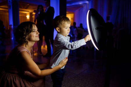 A woman kneels beside a boy pointing at a circular screen with illuminated edges in a dimly lit room.