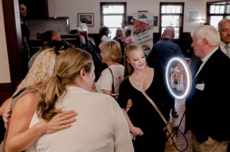 A group of people socialize indoors, with one person using a ring light to take a photo. Background includes various attendees engaging.