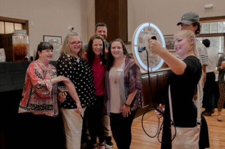 A group of people pose for a photo around a circular light, with one person holding a phone to take the picture.